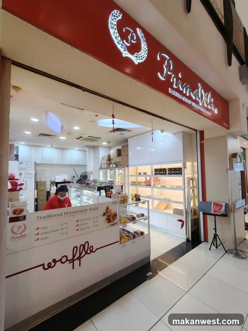 A view of the PrimaDeli bakery storefront. The shop has a red awning with the PrimaDeli logo and name in white illuminated lettering. Below the awning is the entrance to the shop, with a counter displaying waffles and other baked goods. A staff member in a red uniform is visible behind the counter. The interior of the shop features white shelves stocked with products.