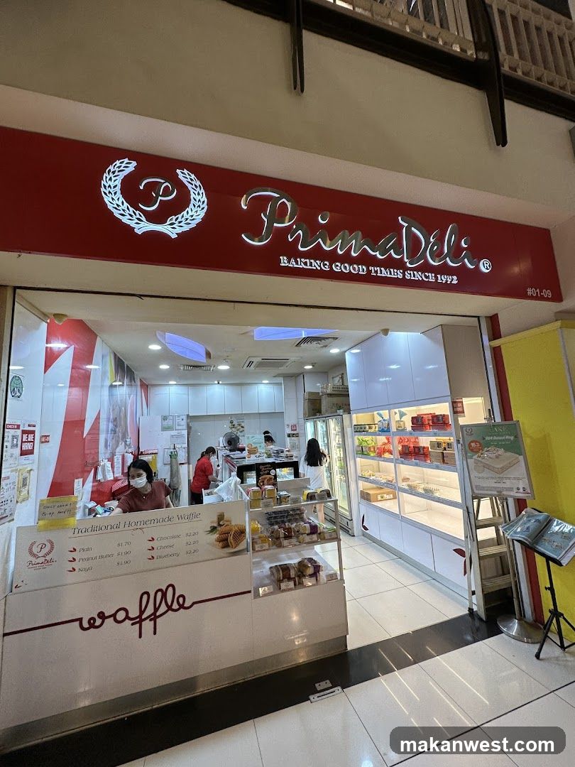 The exterior of a PrimaDeli shop. The shop has a red facade with the PrimaDeli logo and name prominently displayed. Inside, staff members are visible behind a counter, and there are display cases filled with baked goods. A menu board for traditional homemade waffles is also visible.