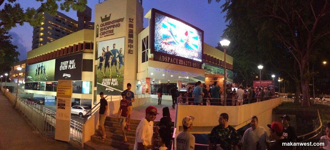 Nighttime exterior view of Queensway Shopping Centre in Singapore, featuring illuminated signage including 'Every Day Every Play' ads and a large digital screen displaying abstract visuals. People are gathered near the entrance on steps and walkways.