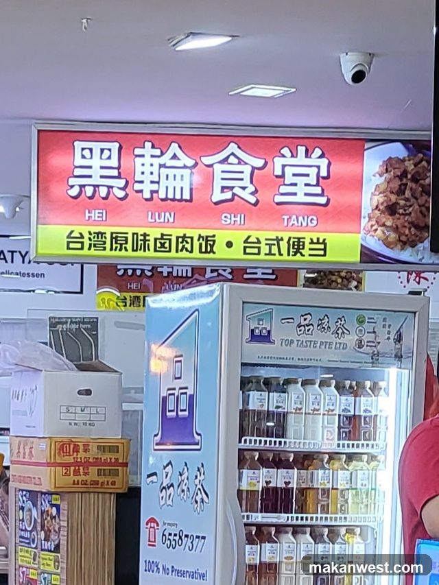 A storefront with a red signboard displaying '黑輪食堂' (HEI LUN SHI TANG) and Chinese characters indicating Taiwanese-style braised pork rice and Taiwanese bento boxes. Below the main sign, there is a yellow banner advertising these items. To the right of the sign, an image of cooked meat is visible. In front of the shop, there are stacked cardboard boxes on the left and a refrigerator filled with bottled beverages labeled '一品冰茶' (Yi Pin Bing Cha). A person in a pink shirt is partially visible on the far right.