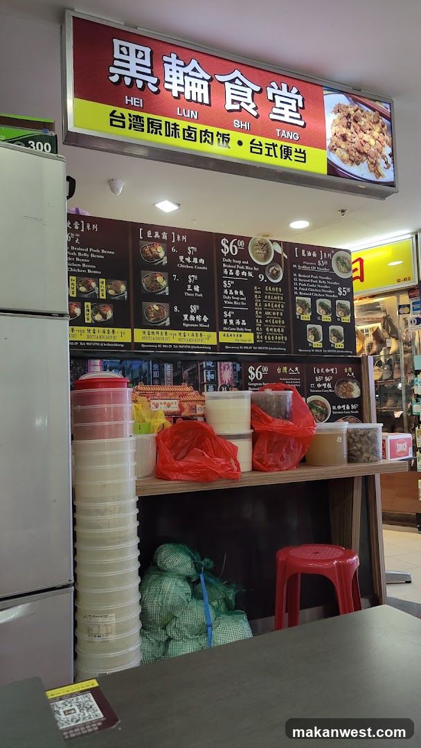 A food stall with a red and yellow signboard displaying '黑輪食堂' (Hei Lun Si Tang) and advertising Taiwanese-style braised pork buns and rice boxes. Below the sign, there is a menu board listing various items like Braised Pork Buns, Chicken Rice, and other dishes with prices in dollars. The counter area has stacked containers, plastic bags, and green-wrapped goods underneath.