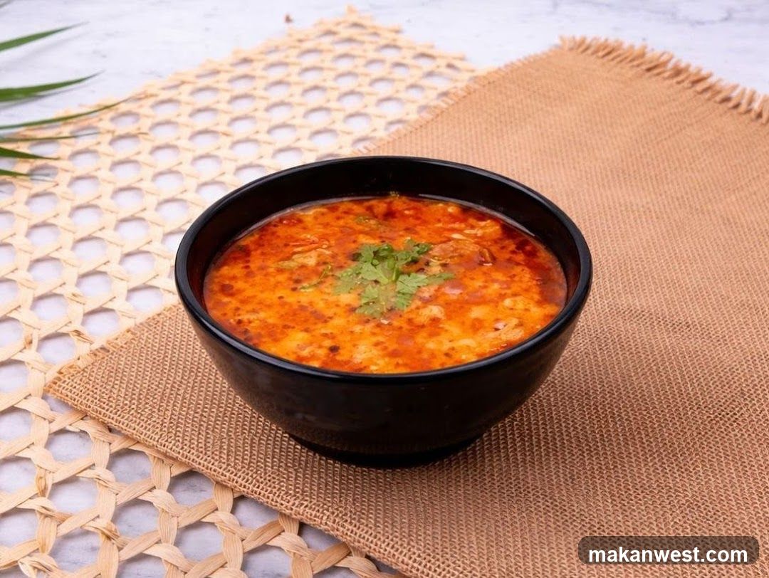A black bowl filled with a vibrant orange-red soup, garnished with fresh green cilantro, sits on a woven placemat. The placemat is placed on a textured surface, with a hint of a green leaf in the upper left corner.