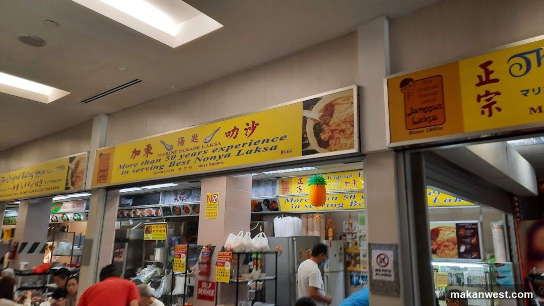 A food stall with yellow signage advertising "Marine Parade Laksa" and "More than 50 years experience in serving Best Nonya Laksa". Another stall to the right advertises "The Original Katong Laksa Janggut Laksa Since 1960s". The stalls are located in a food court or hawker center.