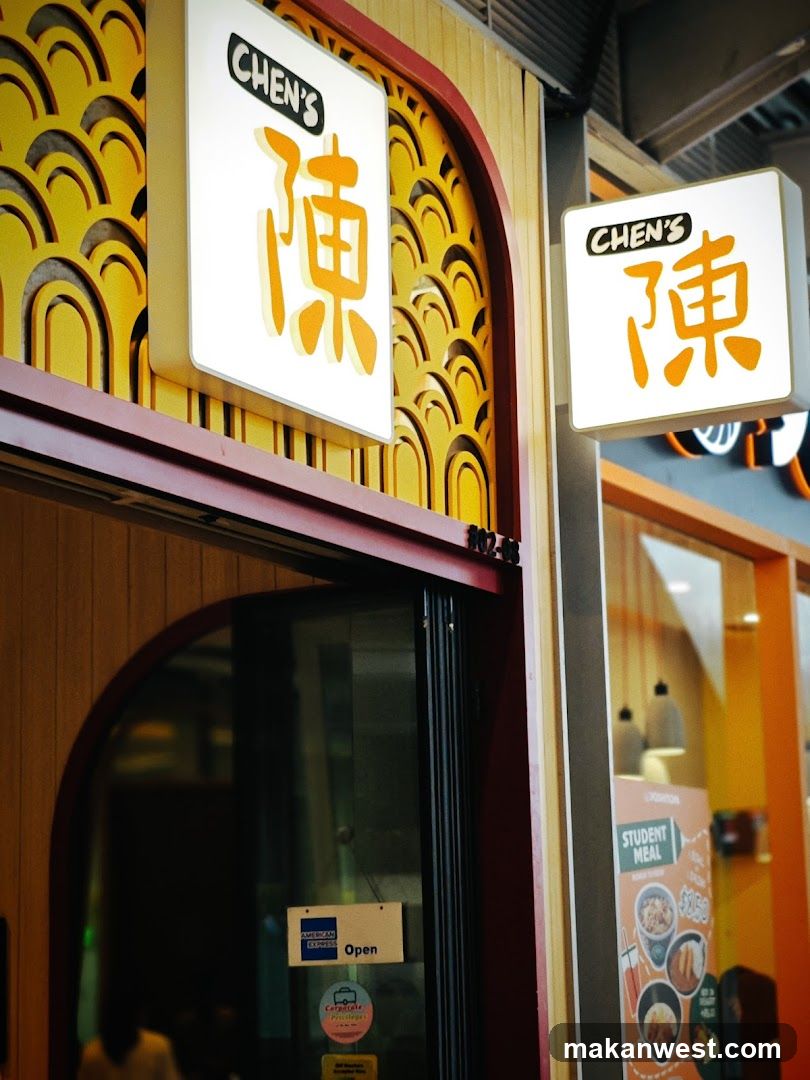 The exterior of a restaurant with two illuminated signs displaying the name "CHEN'S" and Chinese characters. The signs are mounted on a wall with a decorative yellow pattern. Below the signs, a dark-colored doorway with glass panels is visible. Stickers, including an "American Express" logo and an "Open" sign, are affixed to the glass. To the right, a poster advertising a "Student Meal" for $8.50 is displayed in a window.