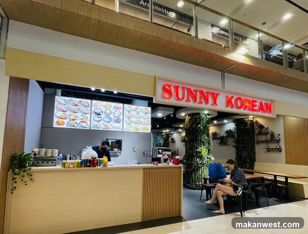 A storefront for a Korean restaurant with a prominent red neon sign that reads "SUNNY KOREAN". There are illuminated menu boards displaying various Korean dishes. A person is working behind the counter, and another person is seated at a table in the dining area. The interior features decorative plants and wooden accents.
