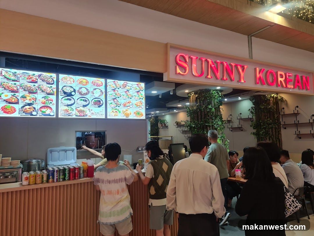 A brightly lit storefront with a large sign reading "SUNNY KOREAN". Below the sign is a menu board displaying various Korean dishes with pictures. Several customers are standing at the counter, and some are seated at tables in the background. The interior features decorative trees and shelves on the walls.