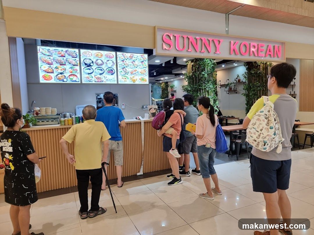 A line of people are waiting to order at a Korean restaurant. The restaurant has a bright pink sign that says "SUNNY KOREAN". There are menu boards displayed above the counter with pictures of food. The interior is decorated with plants.