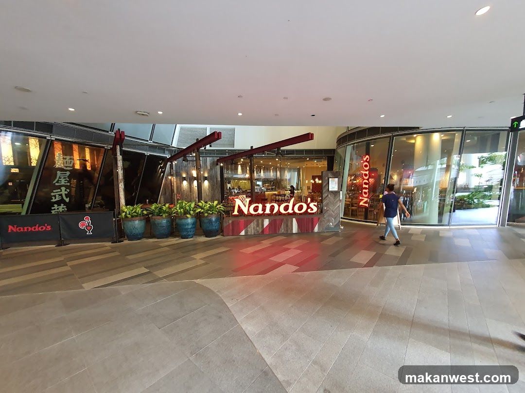 The exterior of a Nando's restaurant with its distinctive red and white signage. The restaurant has large glass windows and a wooden facade. There are several potted plants in front of the restaurant. A person is walking past the entrance.