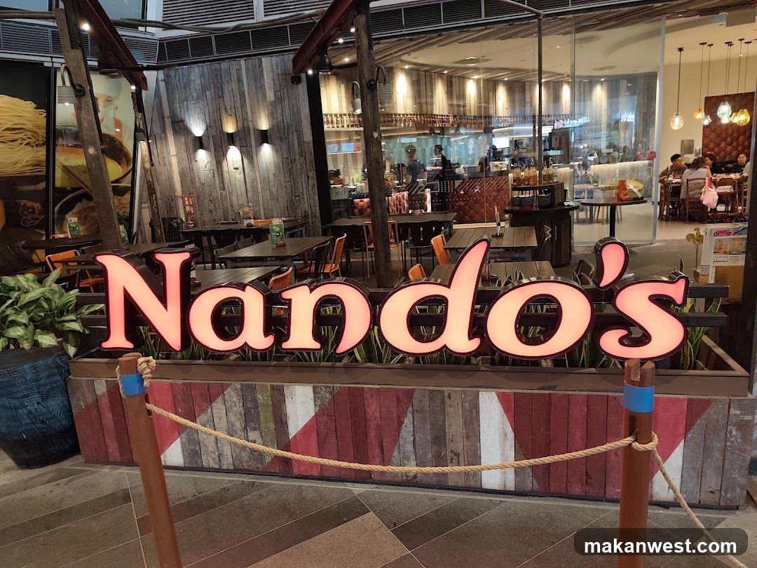 The exterior of a Nando's restaurant with large, illuminated signage. The restaurant interior is visible through the glass facade, showing tables, chairs, and patrons dining. The decor features wooden elements and industrial-style lighting.