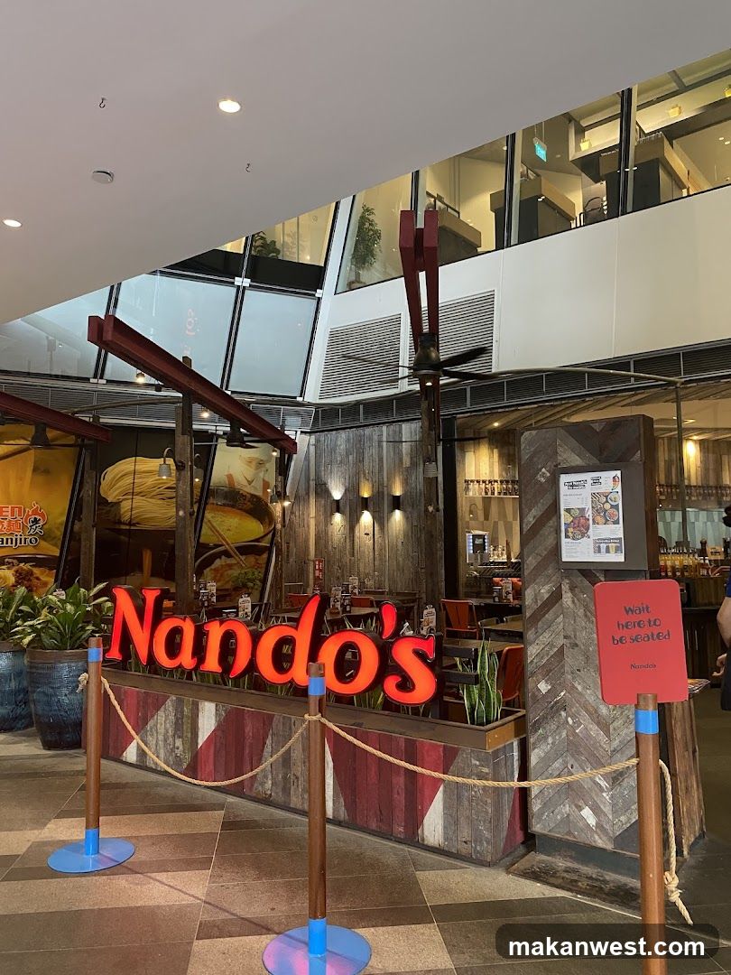 The exterior of a Nando's restaurant with its prominent red neon sign. The entrance is cordoned off with ropes and posts, and a sign indicates 'Wait here to be seated'. The restaurant features a rustic wooden facade and indoor seating is visible through the open entrance.