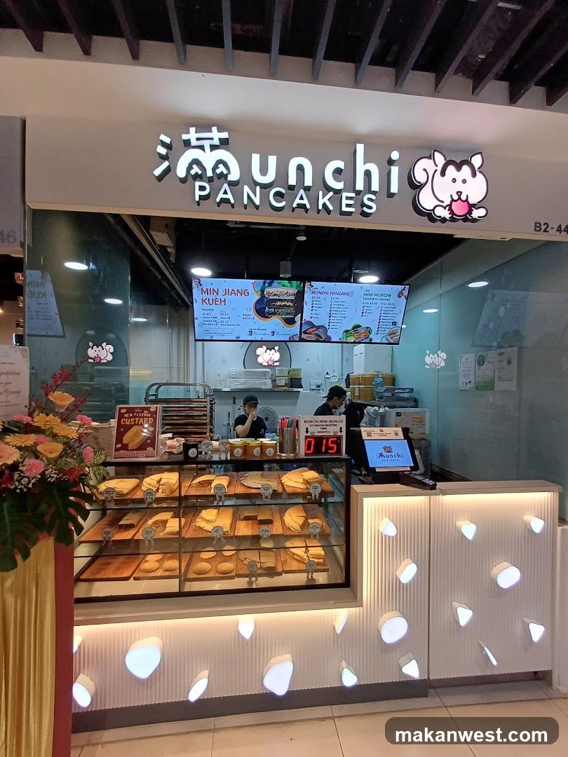 A brightly lit shop front with a display case filled with various pancakes and pastries. The shop's signage features stylized Chinese characters and the name "Munchi PANCAKES" with a cartoon squirrel logo. Behind the counter, staff members are visible, and a digital display shows "015". The interior has a modern design with illuminated decorative elements on the counter.