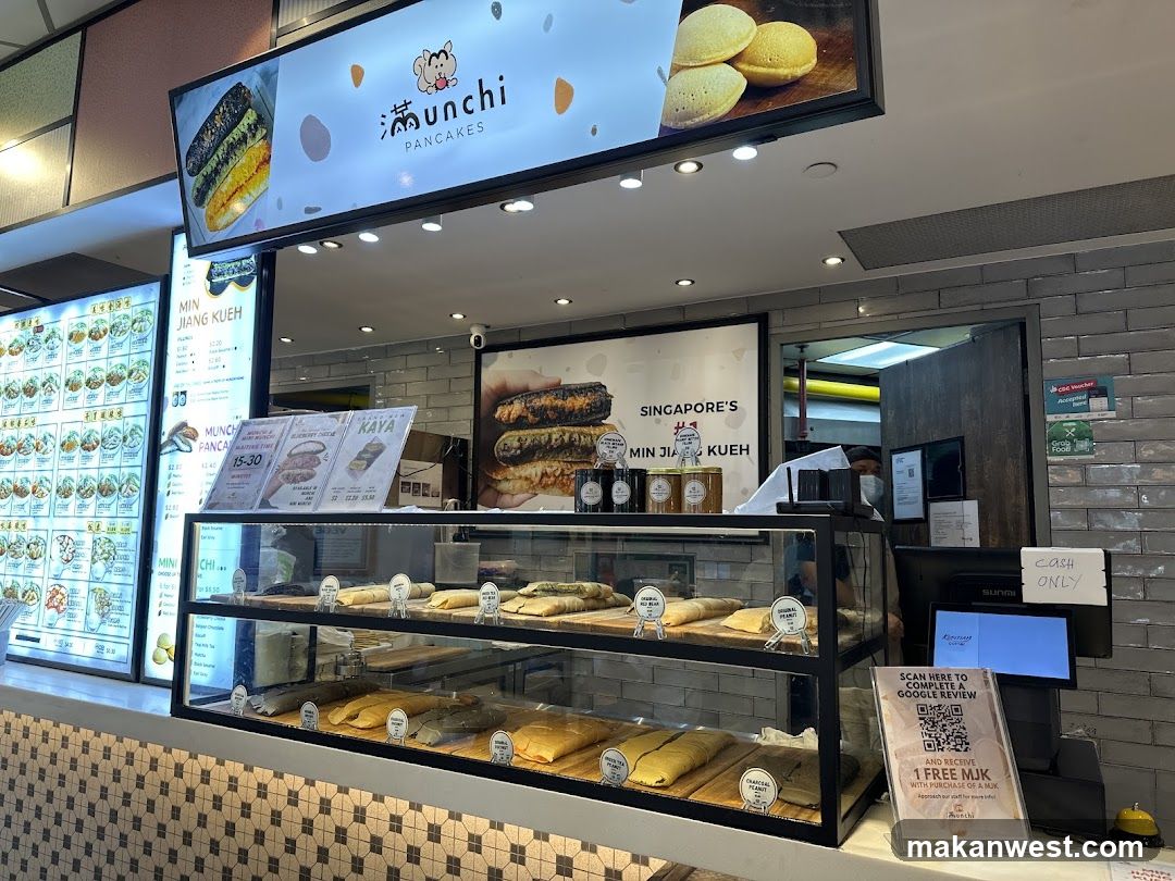 A shop front with a large illuminated sign displaying the business name and images of pancakes. Below the sign is a display case filled with various types of pancakes. To the left, a menu board shows different food items and prices. A cashier counter with a computer monitor and a QR code for a Google review is visible on the right.
