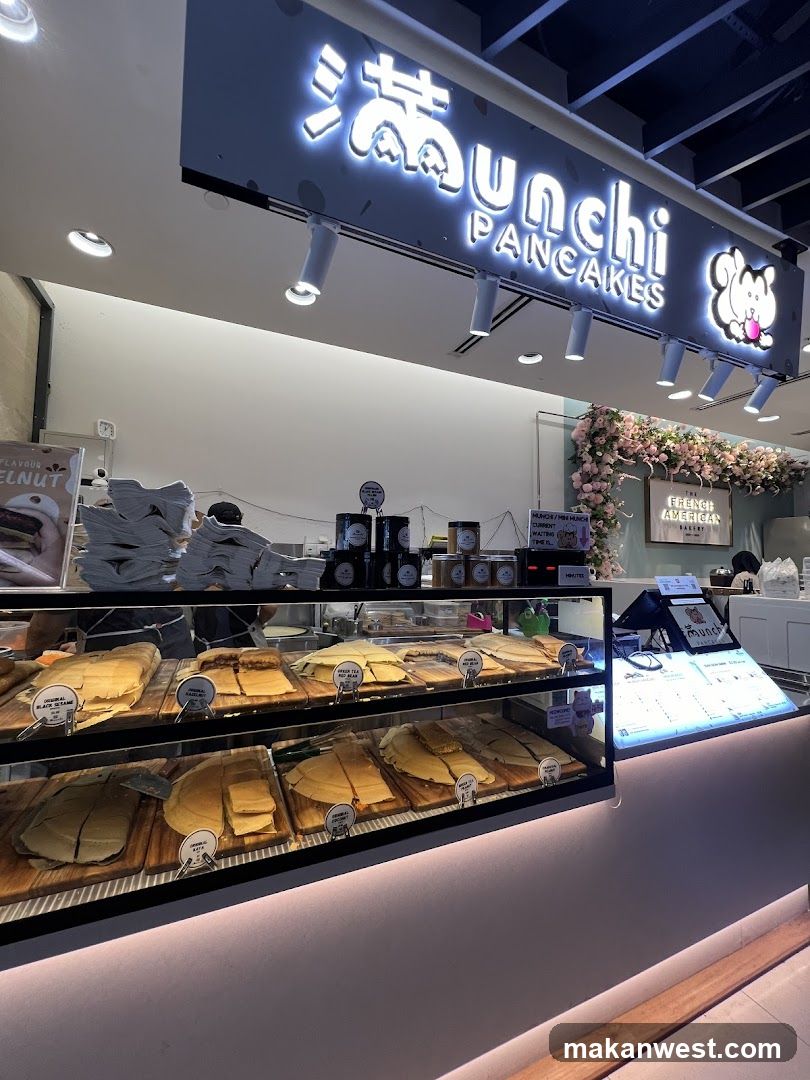 A brightly lit shop front with a prominent sign reading "Munchi PANCAKES" and a cartoon character. Display cases showcase a variety of pancakes, some labeled with flavors like "Original Black Sesame," "Original Kaya," and "Spiced Tea Red Bean." A menu board is visible on the counter.