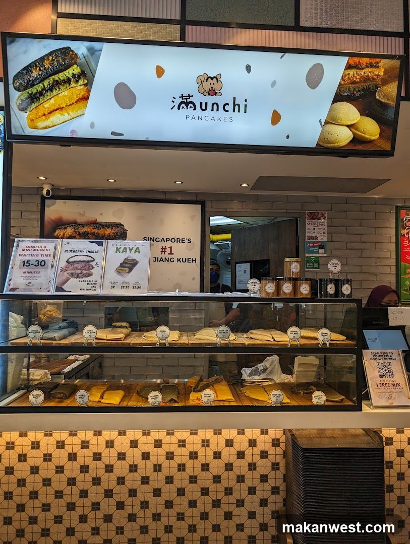 A brightly lit shop front displays a variety of pancakes and related items. A large illuminated sign above features the brand name "Munchi PANCAKES" with images of different pancake varieties. Below the sign, a display case showcases rows of prepared pancakes. A menu board to the left lists "Munchi & Mini Munchi" with a waiting time of "15-30 MINUTES" and highlights flavors like "BLUEBERRY CHEESE" and "KAYA". Another section of the display advertises "SINGAPORE'S #1 JIANG KUEH". The display case itself holds numerous pancakes, with labels indicating flavors such as "ORIGINAL PEANUT", "GREEN TEA PEANUT", and "CHARCOAL PEANUT". A cashier counter is visible on the right with a sign indicating "CASH ONLY" and a QR code for a Google review.