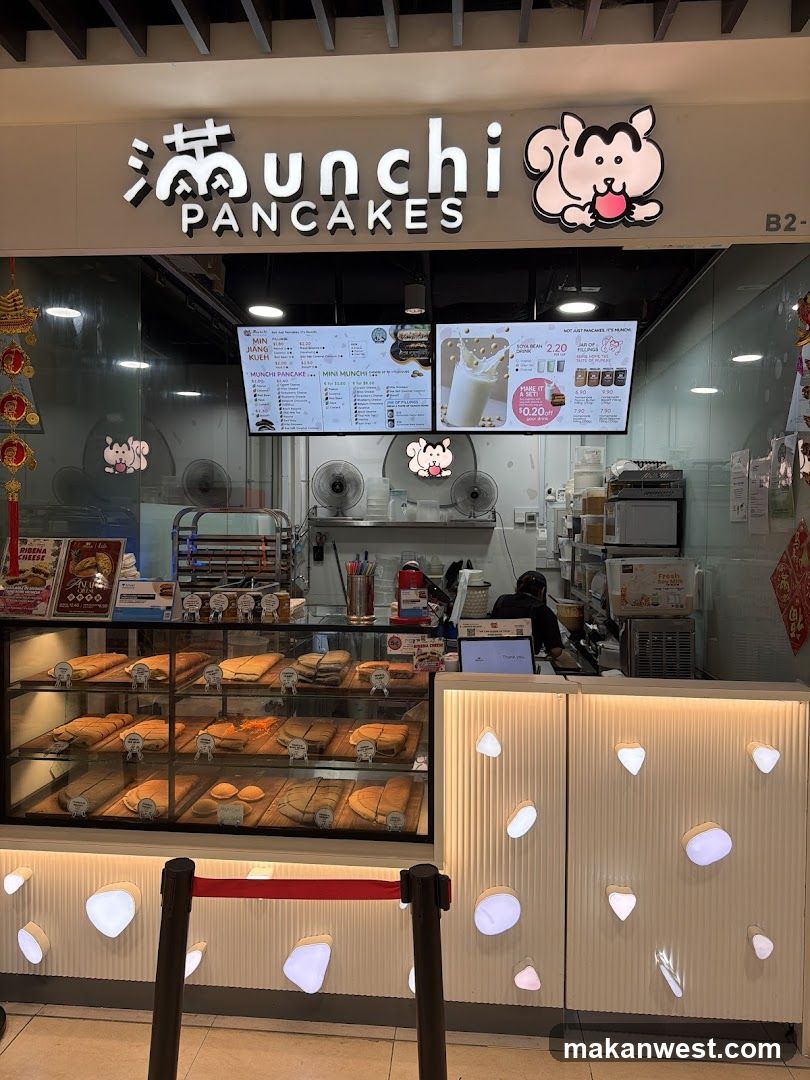 A brightly lit pancake shop with a prominent sign featuring the business name and a cartoon character. Display cases showcase various filled pancakes. Digital menu boards are visible above the counter, and the counter area has decorative lighting.