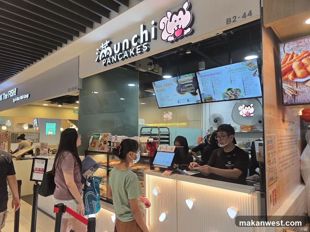 A storefront for a pancake shop with a sign that says "Munchi PANCAKES". There are digital menus displaying various pancake and kueh options. Staff members are visible behind the counter, and customers are queuing. The shop is located in a mall.