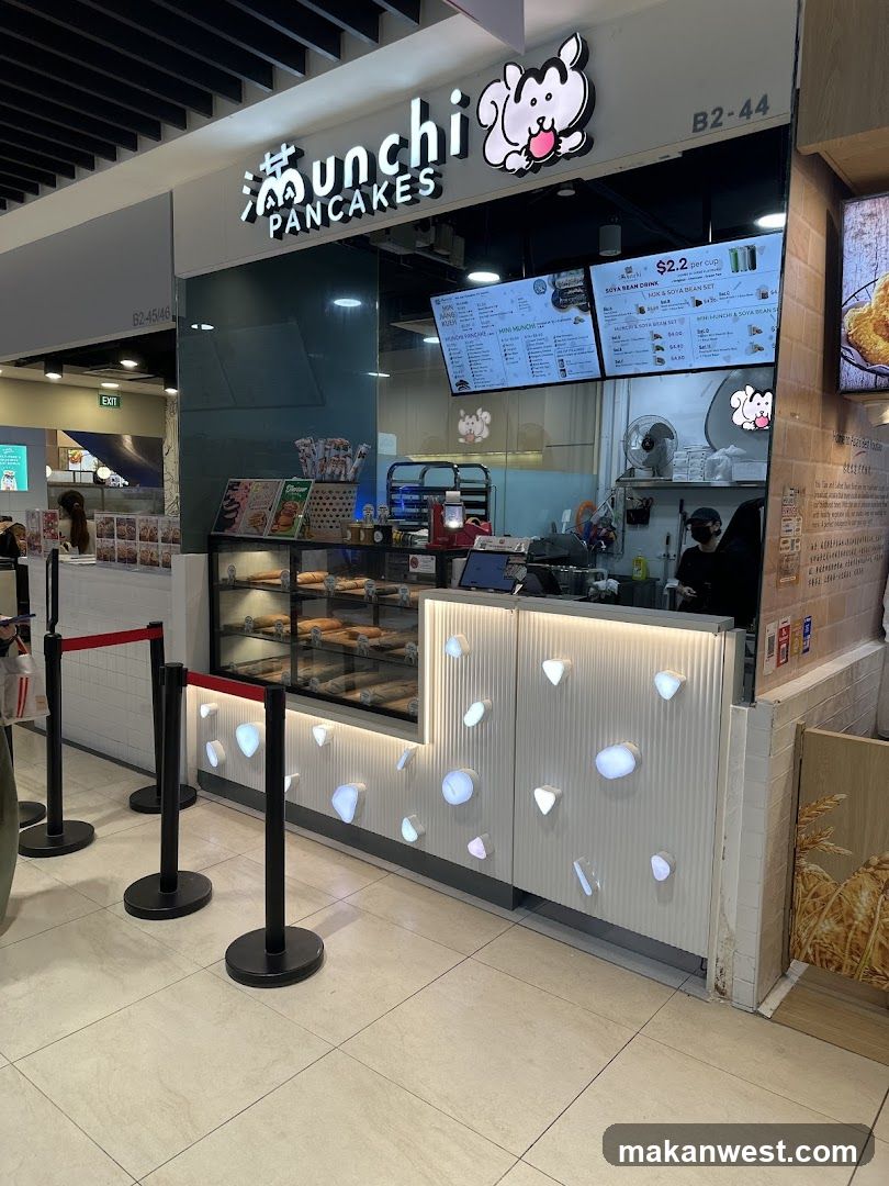 A brightly lit pancake shop with a prominent sign featuring a cartoon character. The storefront displays a glass case filled with various pancakes and a menu board above the counter. Red velvet ropes guide customers.