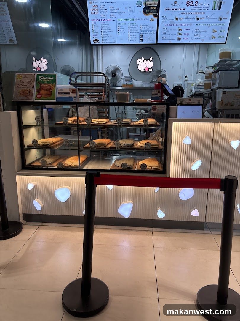 A food stall with a display case filled with pastries and a digital menu board above. The stall has a modern design with illuminated decorative elements on the wall. A red velvet rope barrier is in the foreground.