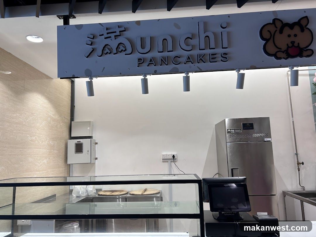 A sign with stylized text and a cartoon character is mounted above a counter. The text appears to read "Unchi PANCAKES". To the right of the text is a cartoon drawing of a brown, dog-like creature with a red ball in its mouth. Below the sign, there are several spotlights. In the background, there is a stainless steel refrigerator and a cash register. A glass display case is in the foreground.