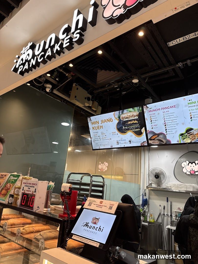 A view of the Munchi Pancakes shop front, showing the illuminated sign, menu boards, display cases with food, and a digital ordering screen.