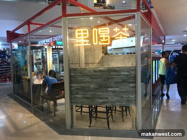 A glass-enclosed shop front with a red frame. Inside, people are seated at tables. A neon sign with Chinese characters is visible above the counter. The exterior also shows reflections of the surrounding mall environment.
