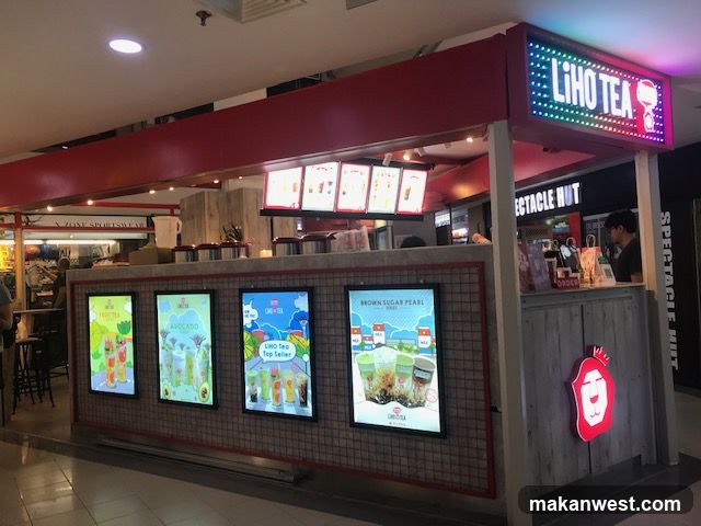 A brightly lit Liho Tea kiosk is shown in a mall. The kiosk has a red awning and a sign with the Liho Tea logo and name in colorful LED lights. Below the sign, there are illuminated posters displaying various drinks, including fruit tea, avocado drinks, and brown sugar pearl series. A staff member is visible behind the counter.