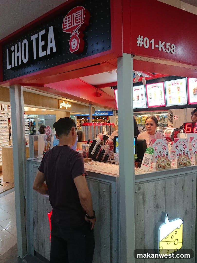 A Liho Tea shop front with a prominent sign displaying the brand name. A customer is standing at the counter, and staff members are visible behind it. The shop has a modern design with a concrete-like texture on the lower half and a red upper section. There are menu boards with pictures of drinks displayed.