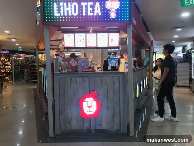 A Liho Tea kiosk with a prominent LED sign displaying the brand name. The kiosk has a wooden facade with a large red lion logo. There are menu boards with pictures of drinks and prices, and a digital display showing '98'. Two customers are standing at the counter, looking at the menu. The kiosk is located in a shopping mall with other shops visible in the background.