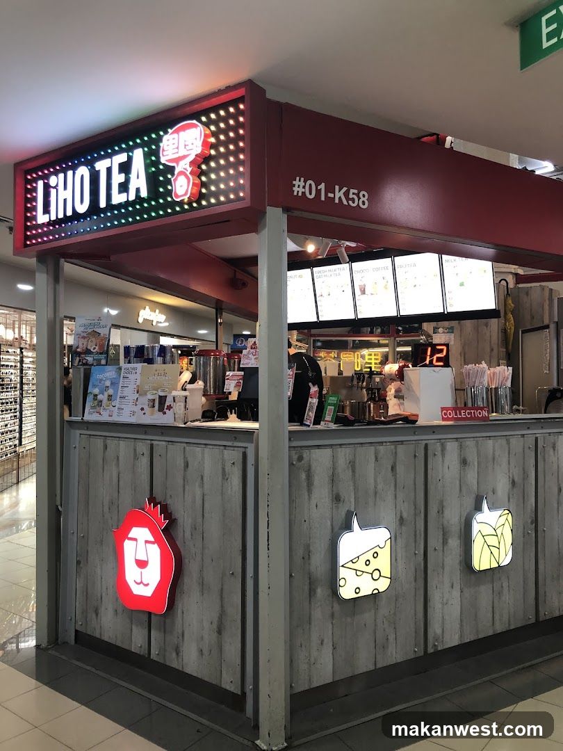 A red and grey kiosk with a prominent "LIHO TEA" sign illuminated by colorful LED lights. The kiosk has a wooden-textured facade decorated with illuminated icons of a lion's head, a slice of cheese, and a leaf. Behind the counter, menus are displayed on screens, showing various drink options. The unit number "#01-K58" is visible above the counter.