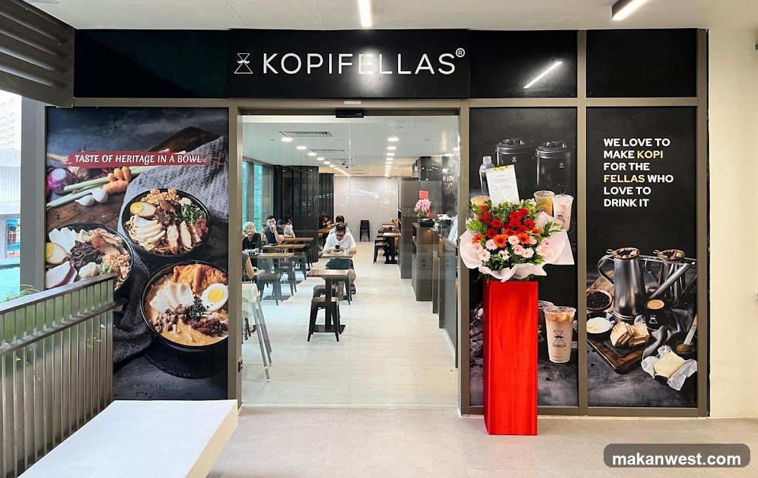 The storefront of a cafe with large graphics on the windows. One graphic displays three bowls of food with the text "TASTE OF HERITAGE IN A BOWL". Another graphic shows coffee drinks and text that reads "WE LOVE TO MAKE KOPI FOR THE FELLAS WHO LOVE TO DRINK IT". Inside, tables and chairs are visible with a few customers seated. A floral arrangement in a red vase stands outside the entrance.