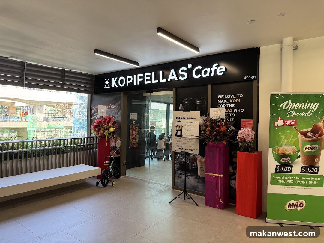 The storefront of Kopifellas Cafe features a prominent sign with the cafe's name. To the right of the entrance, a large banner advertises an "Opening Special" for Milo drinks, with prices listed for hot and iced versions. Inside the cafe, patrons can be seen seated at tables. Outside, there are decorative floral arrangements and a long white bench. A small bicycle is parked near the entrance. Construction is visible in the background through a railing.