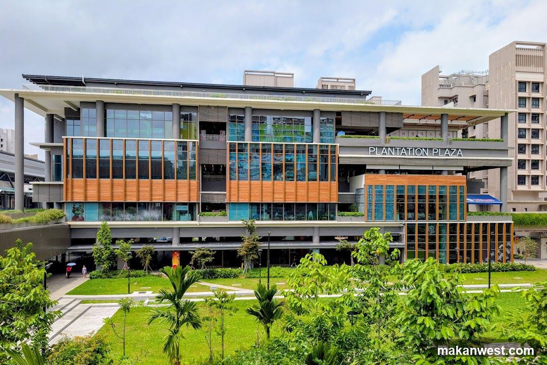 A modern multi-story building with a facade of glass and wood-toned panels. The building has multiple levels with large windows and balconies. A prominent sign reads "PLANTATION PLAZA". Greenery and trees surround the building, with a paved walkway and grassy areas in the foreground.