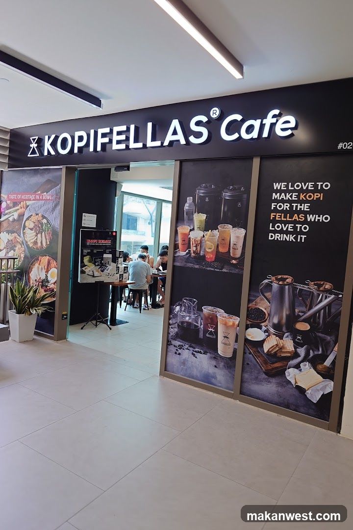 The storefront of KOPIFELLAS Cafe features a large illuminated sign with the cafe's name. To the left, a poster displays images of various noodle dishes with the text "TASTE OF HERITAGE IN A BOWL". To the right, a large graphic showcases an assortment of iced beverages and a coffee brewing setup with toast and butter, accompanied by the text "WE LOVE TO MAKE KOPI FOR THE FELLAS WHO LOVE TO DRINK IT". A menu board is visible near the entrance. Inside, patrons are seated at tables.