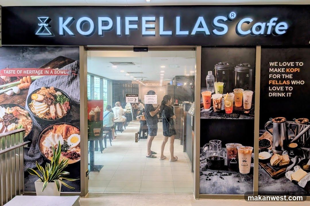 The storefront of Kopifellas Cafe features a large illuminated sign with the cafe's name. The exterior is decorated with large posters showcasing food and drinks. On the left, posters display bowls of noodles and other dishes with the text "TASTE OF HERITAGE IN A BOWL". On the right, posters show various colorful beverages in plastic cups and a display of coffee-making equipment with the text "WE LOVE TO MAKE KOPI FOR THE FELLAS WHO LOVE TO DRINK IT". The entrance is a glass door leading into the cafe, where customers can be seen seated at tables.