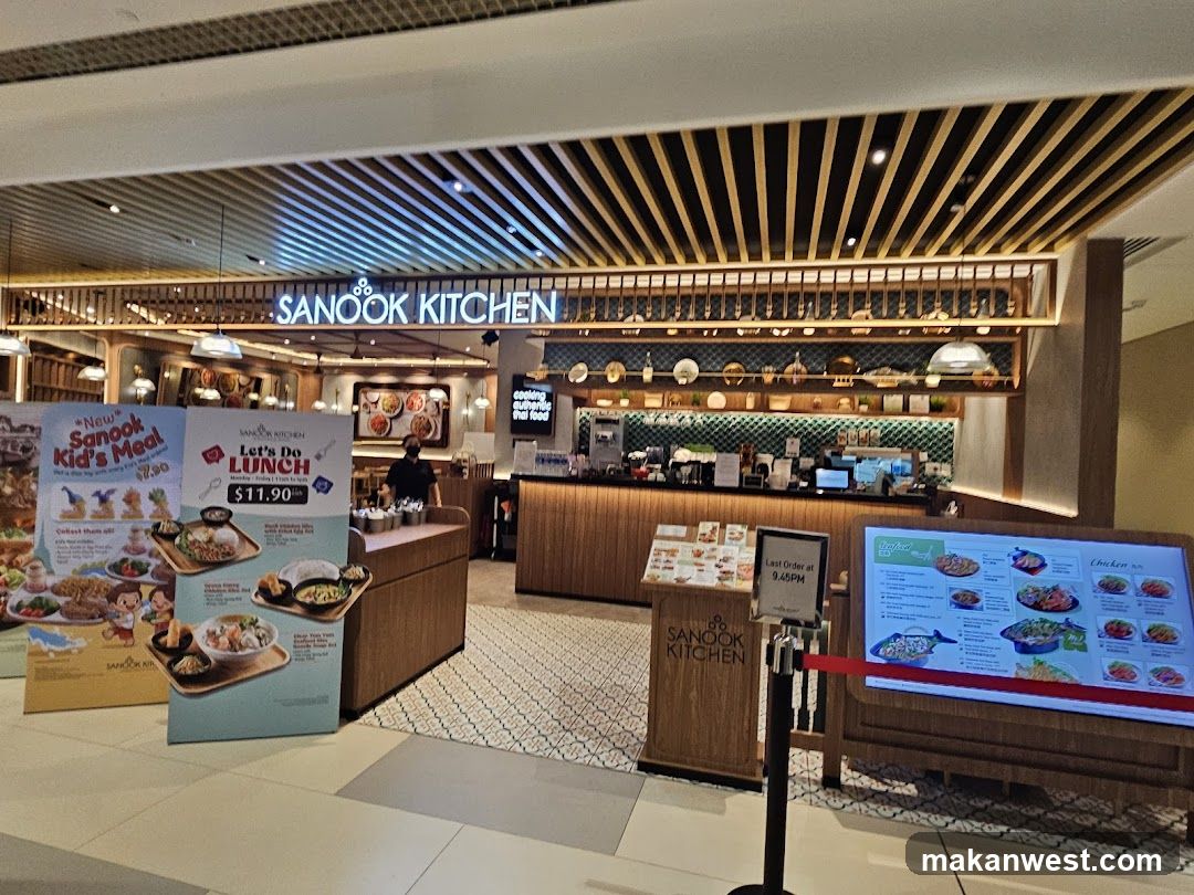 The exterior of a restaurant called Sanook Kitchen. There are promotional posters for "New Sanook Kid's Meal" and "Let's Do Lunch". A digital menu board displays various food items. The restaurant interior is visible behind the counter, with shelves displaying decorative items.