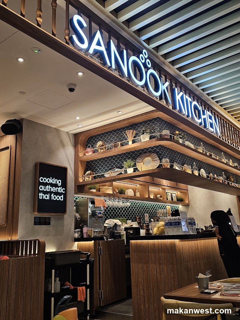A neon sign that reads "SANOOOK KITCHEN" is prominently displayed above the entrance to a restaurant. Below the sign, shelves are stocked with decorative items and kitchenware. A sign on the wall states "cooking authentic thai food." The interior features wooden accents and a counter area where a person is visible.