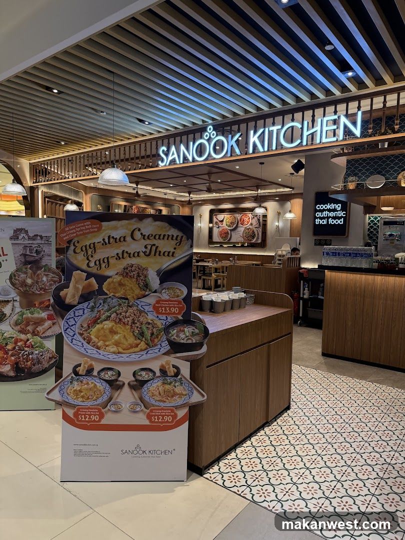 A brightly lit restaurant interior with a prominent sign reading "SANOOK KITCHEN". A display stand in the foreground features images of Thai food dishes with prices. The restaurant appears to specialize in authentic Thai cuisine, as indicated by a sign that says "cooking authentic thai food". The flooring has a decorative tile pattern.
