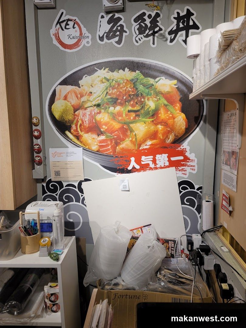 A large poster on a wall displays a bowl of Kaisen Don, a Japanese rice bowl dish with raw fish. The poster has Japanese characters and the logo 'KEI KaisenDon'. Below the bowl, red text reads '人气第一' (Popularity First). To the left of the poster are three small Daruma dolls. To the right, shelves hold stacks of paper cups and chopsticks. The lower part of the image shows various items on shelves and a desk, including pens, a digital clock, plastic containers, and electrical outlets.