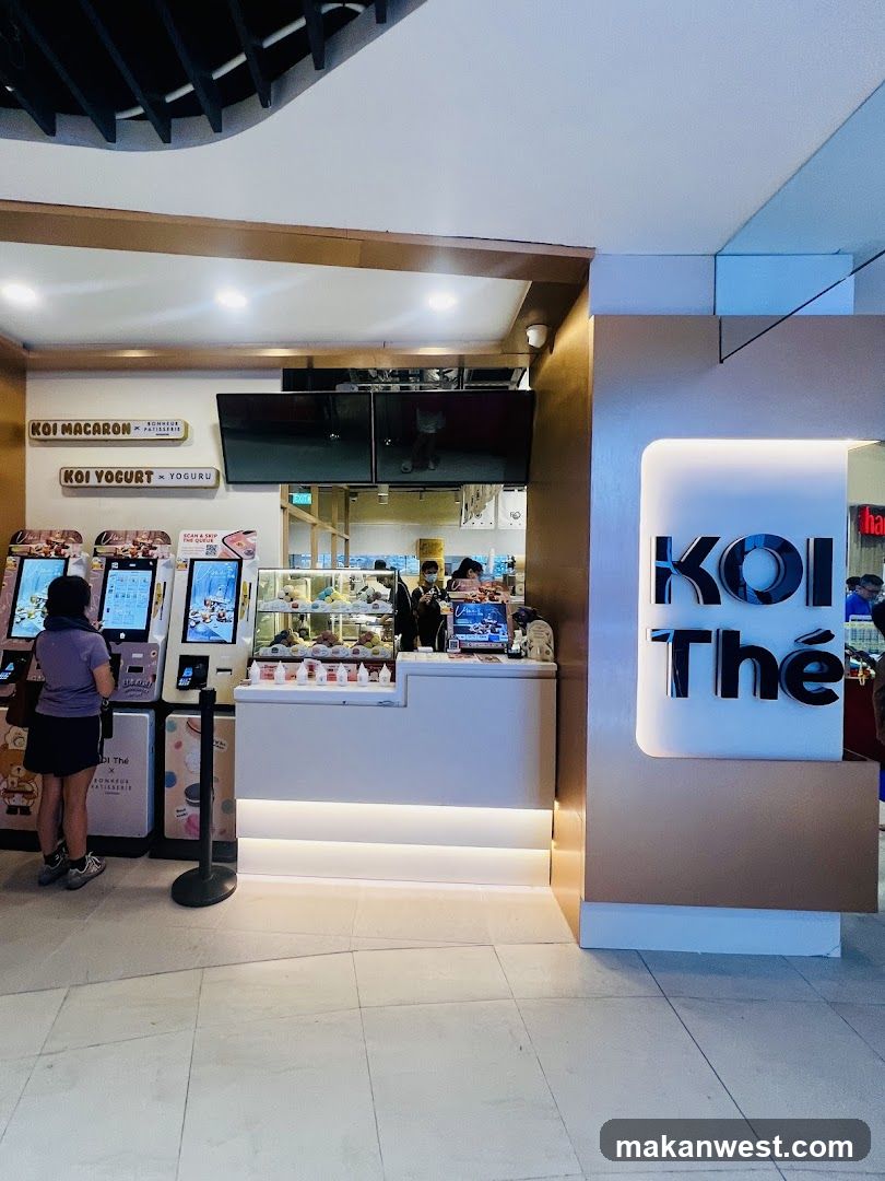 A modern shop front with a prominent illuminated sign that reads "KOI Thé". To the left, there are self-order kiosks and a counter displaying pastries and drinks. Signs above indicate "KOI MACARON x BONHEUR PATISSERIE" and "KOI YOGURT x YOGURU".