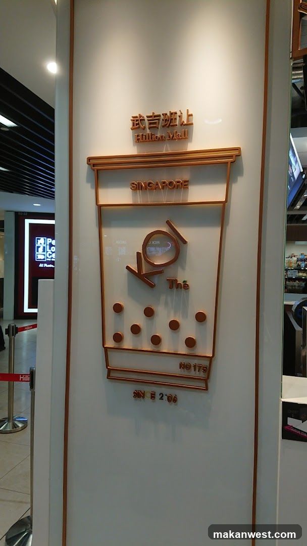 A 3D sign shaped like a cup of bubble tea is mounted on a white wall. The sign has copper-colored lettering and accents. At the top, it reads "武吉班让 Hilton Mall". Below that, "SINGAPORE" is displayed. The central part of the sign features the logo "KOI Thé" with a circular element inside the 'O'. Below the logo, several brown circles represent tapioca pearls. At the bottom, it says "NO 175" and "SINCE 2006". To the left, a red stanchion with a white rope and a sign that says "Hilli" is visible. In the background, a digital screen displays "Pick Up".