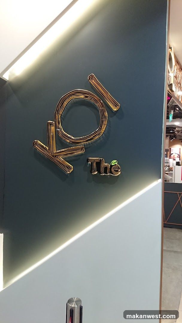 A metallic, gold-colored sign with the text "Koi The" is mounted on a dark teal wall. The letters of "Koi" are stylized and appear to be made of wire. The word "The" is smaller and positioned below and to the right of "Koi," with a small green leaf graphic above the 'e'. The wall is illuminated by a strip of white LED lighting along the top and a diagonal strip of lighting below. To the right, a glimpse of a store interior is visible, with shelving and what appears to be a counter.
