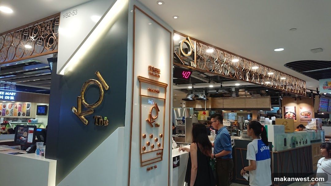 A storefront for a beverage shop with a large golden logo that reads "Koi The". There is a digital display showing "40". People are queuing to order. The shop is located in a mall with other food establishments visible in the background.