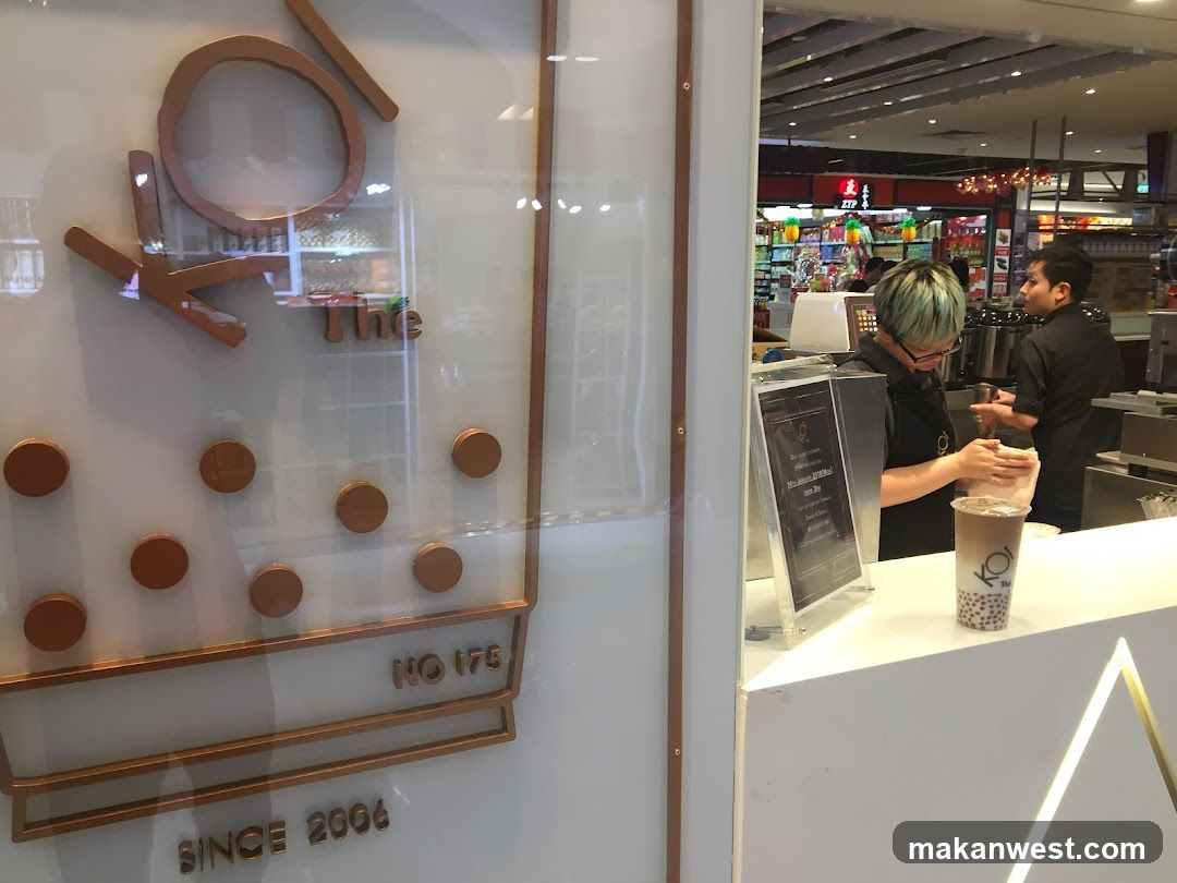 A storefront with a large sign that says "KOI The" and "SINCE 2006". Inside, two staff members are visible behind a counter. One is preparing a drink, and the other is talking to them. A bubble tea drink is on the counter.