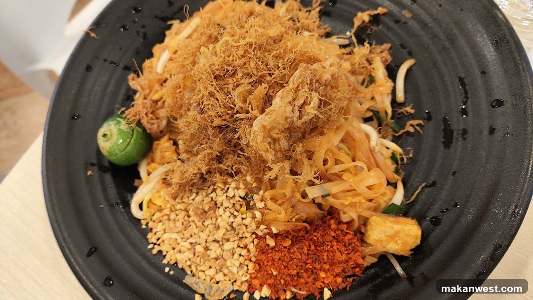 A close-up shot of a plate of Pad Thai, a popular Thai noodle dish. The noodles are stir-fried with bean sprouts, tofu, and a savory sauce. The dish is topped with a generous portion of crispy fried shredded chicken or pork, crushed peanuts, and chili flakes. A lime wedge is placed on the side for added flavor. The black plate has a textured surface and some liquid splatters are visible. The background is blurred, showing a light-colored table and a hint of white furniture.