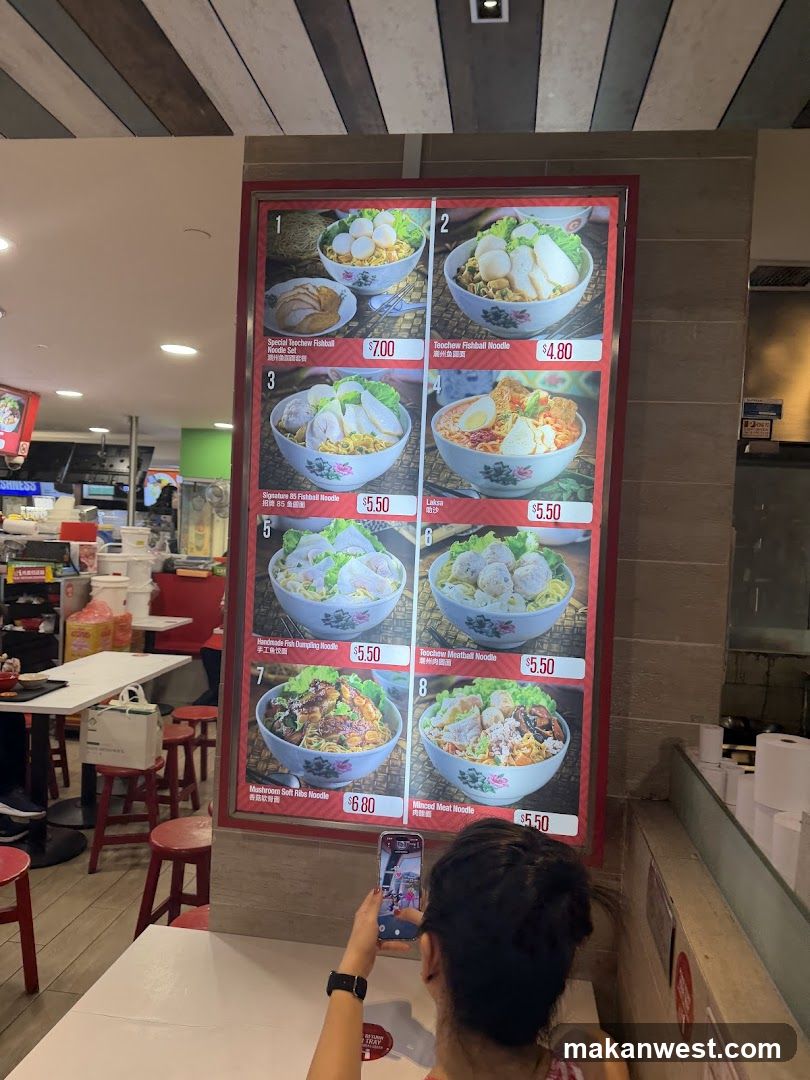 A woman is taking a picture of a menu board with her phone. The menu board displays various noodle dishes with prices.