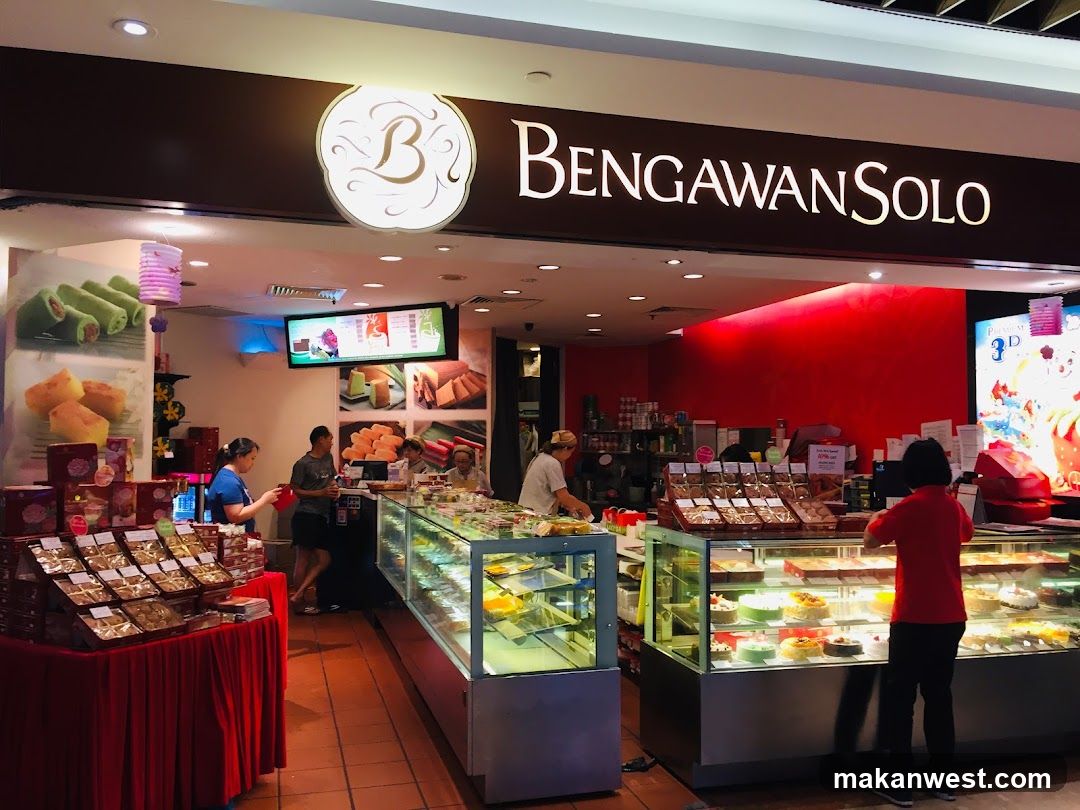 A bakery storefront with a large sign that reads "BENGAWAN SOLO". Inside, display cases are filled with cakes and pastries. Several staff members are visible behind the counter, and customers are browsing the merchandise. The walls are painted red, and there are promotional posters on display.