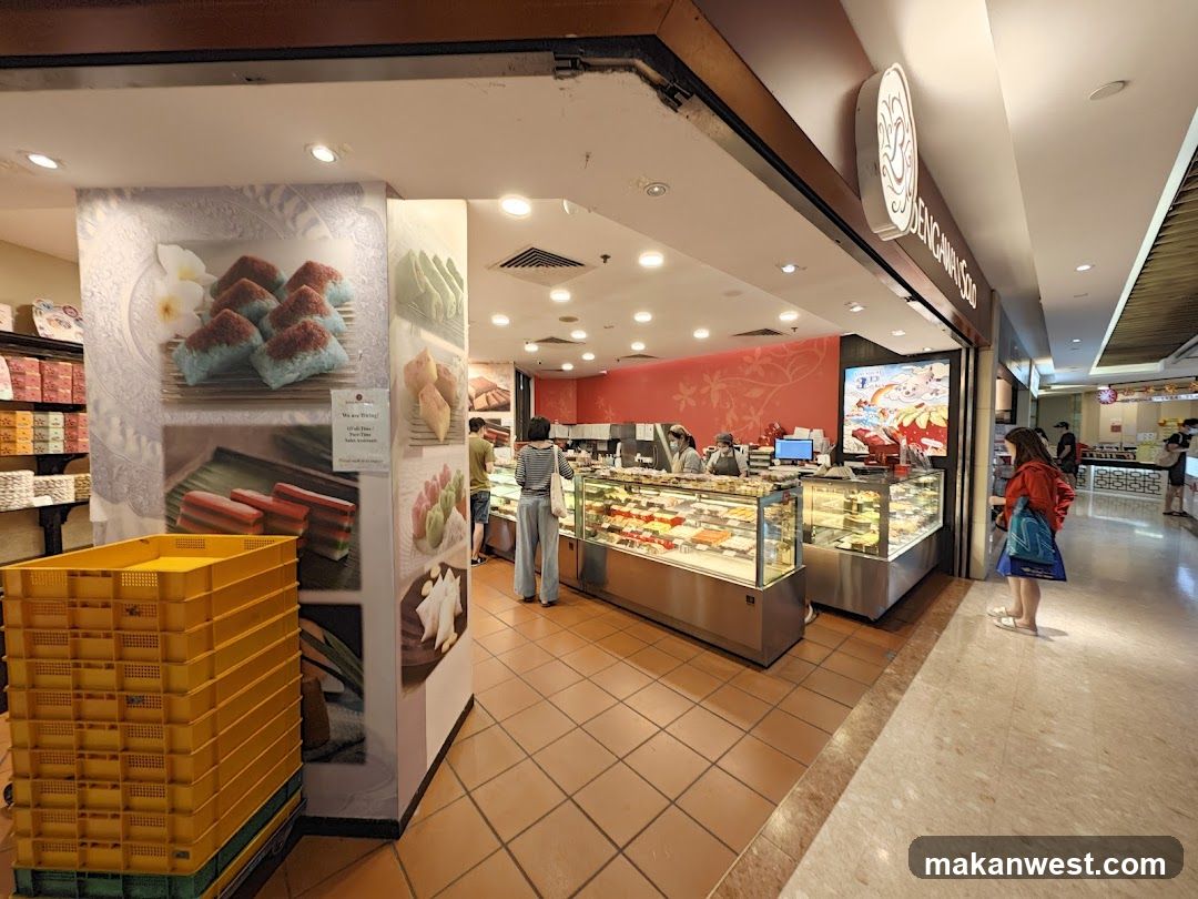 A bakery shop front with a display of cakes and pastries. There are yellow crates stacked to the left and a large mural on the wall depicting various desserts. A woman in a red jacket is standing in front of the display case.