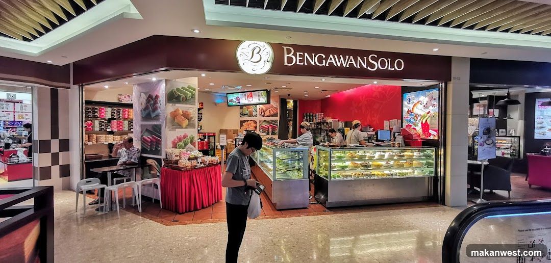 A bakery shop with a display of cakes and pastries. There are staff members serving customers. The shop has a prominent sign that says "BENGAWAN SOLO". There are also images of cakes and pastries displayed on the walls.