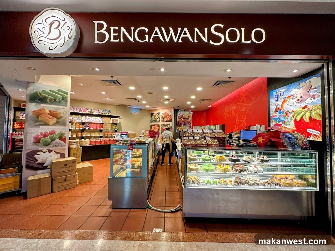A bakery storefront with a large display case filled with cakes and pastries. Shelves behind the counter are stocked with boxed goods. A large banner on the left displays images of various desserts. A colorful advertisement for 3D cakes is on the right.