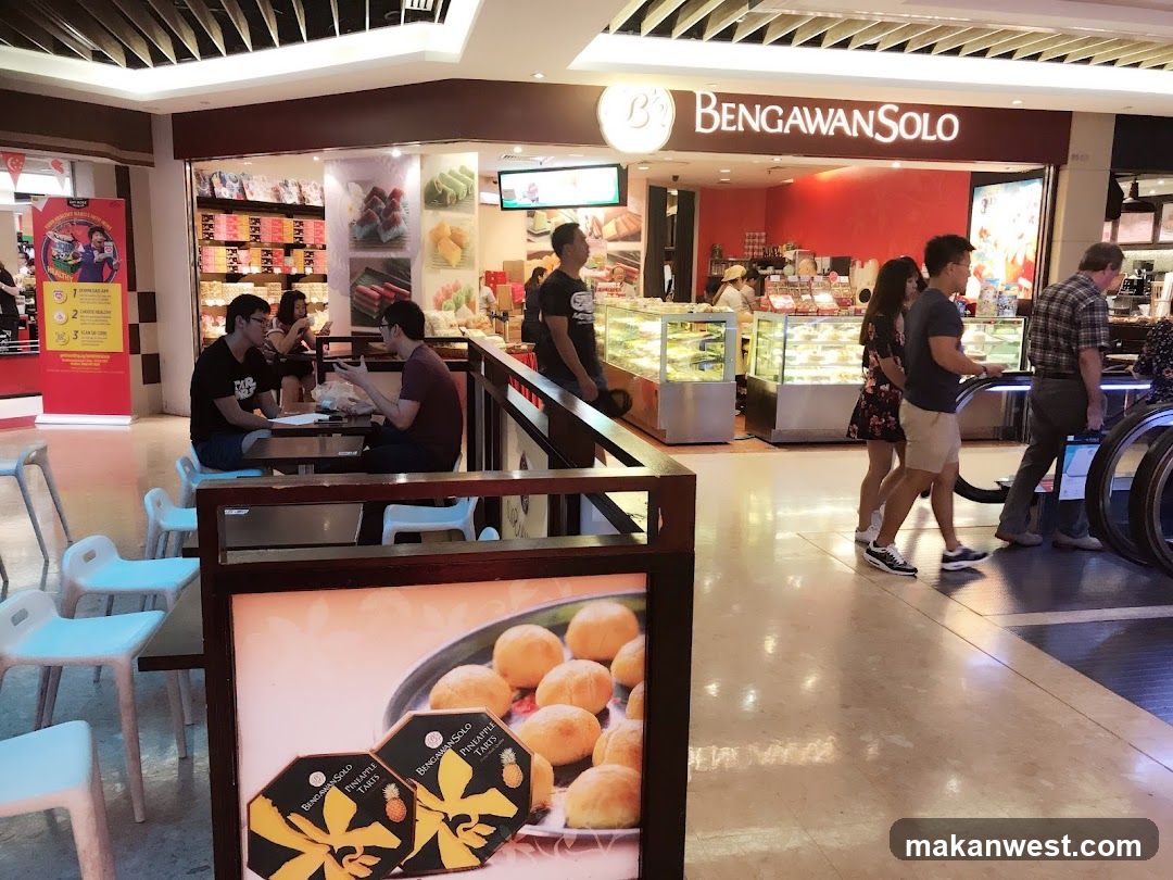 A shop front with a red accent wall and display cases filled with pastries and cakes. People are seen walking past the shop and some are seated at tables outside. A sign for "Bengawan Solo" is visible above the entrance.